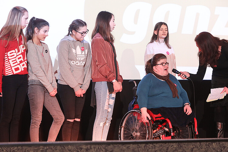 Eine Gruppe von Schülern steht auf der Bühne. Ein Mädchen im Rollstuhl wird interviewt.