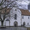 Eine romanische Kirche in Seitenansicht mit heller Fassade, rot eingedecktem Dach und Glockenturm.