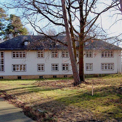 Im Hintergrund ist ein längliches Gebäude zu sehen, vorne eine Rasenfläche und Bäume