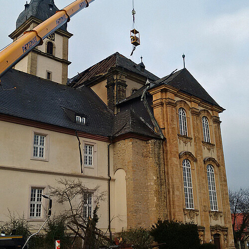 Vor einer Kirchenfassade ist ein Kran-Arm mit einer Gondel ausgefahren. Aus der Gondel werden Teile der beschädigten Dachkonstruktion geborgen.