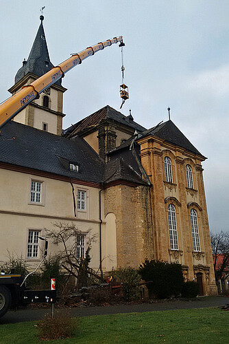 Vor einer Kirchenfassade ist ein Kran-Arm mit einer Gondel ausgefahren. Aus der Gondel werden Teile der beschädigten Dachkonstruktion geborgen.