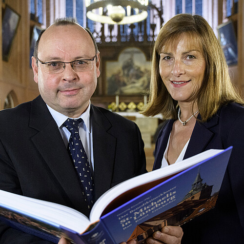 Zwei Personen stehen in einer Kirche und halten ein aufgeschlagenes Buch in den Händen.