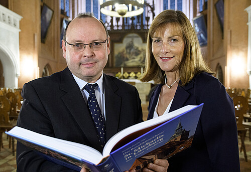 Zwei Personen stehen in einer Kirche und halten ein aufgeschlagenes Buch in den Händen.