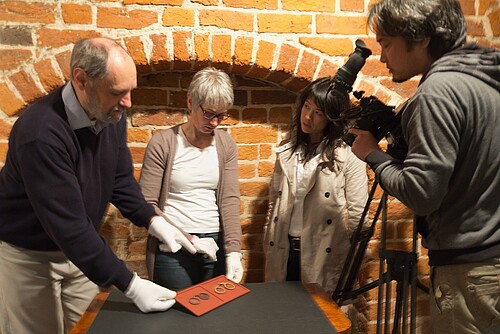 Zwei Mitarbeiter der Klosterkammer, eine japanische Journalistin und ein Kameramann stehen um einen kleinen Tisch herum und schauen gemeinsam auf zwei historische Brillen.