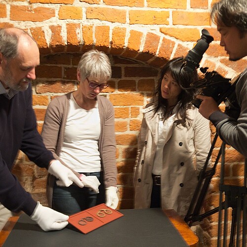 Zwei Mitarbeiter der Klosterkammer, eine japanische Journalistin und ein Kameramann stehen um einen kleinen Tisch herum und schauen gemeinsam auf zwei historische Brillen.