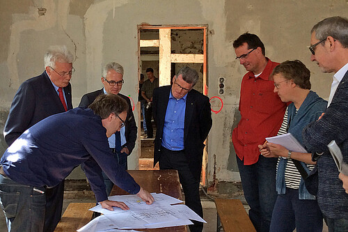 Sieben Personen stehen in einem Gebäude, in dem gebaut wird, um einen hölzernen Tisch auf dem Pläne liegen.