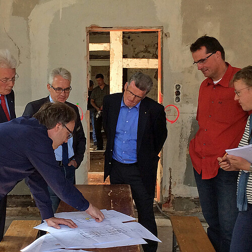 Sieben Personen stehen in einem Gebäude, in dem gebaut wird, um einen hölzernen Tisch auf dem Pläne liegen.