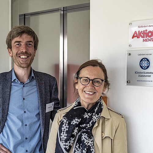 Johannes König, Silke Sackmann vor einer Eingangstür. Schilder Aktion Mensch und Klosterkammer Hannover.