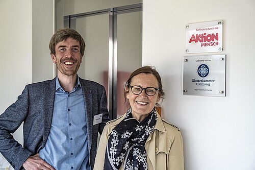 Johannes König, Silke Sackmann vor einer Eingangstür. Schilder Aktion Mensch und Klosterkammer Hannover.