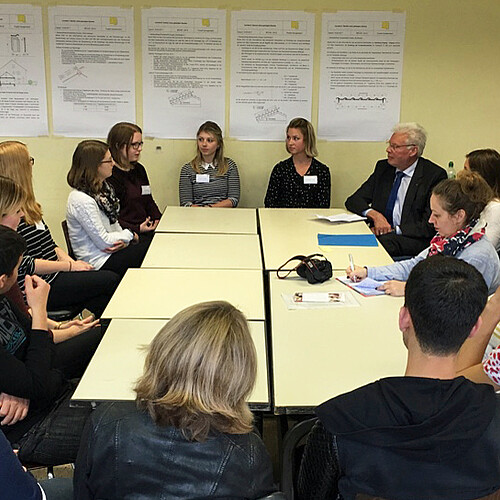 Eine Gruppe von Jugendlichen und Erwachsenen sitzt in einem Klassenraum um einen Tisch herum.