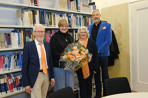 Vier Personen stehen in einer Bibliothek vor Bücherregalen.