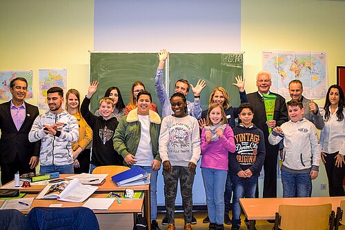 Eine Gruppe von Realschülerinnen und -schülern und Erwachsene stehen in einem Klassenraum.