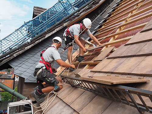Zwei Männer mit Bauhelmen, angeseilt, decken ein Dach. Baugerüst.