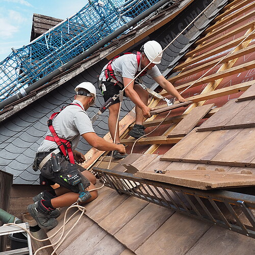Zwei Männer mit Bauhelmen, angeseilt, decken ein Dach. Baugerüst.