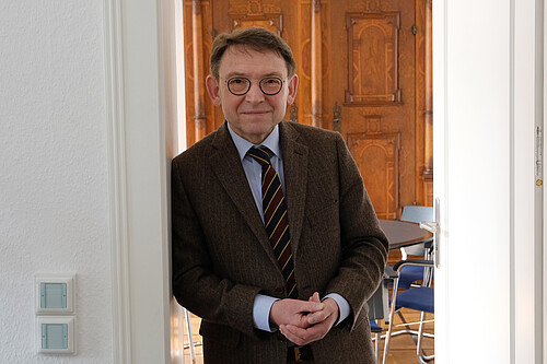Ein Mann in braunem Sakko, mit Krawatte und Brille steht in einem Türrahmen.