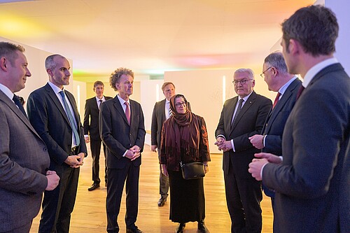 neun Personen stehen in einem Halbkreis in einem Ausstellungsraum, der in hellen Farben gestaltet ist.