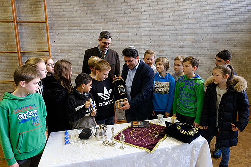 Eine Gruppe Schülerinnen und Schüler und zwei Erwachsene stehen um einen Tisch mit Gegenständen aus dem jüdischen Leben herum.