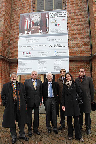 Sechs Personen stehen vor einem Baustellenschild vor einer Kirchenfassade.