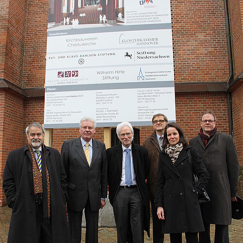 Sechs Personen stehen vor einem Baustellenschild vor einer Kirchenfassade.