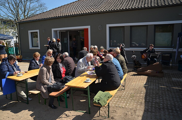 Eine Gruppe von Personen sitzt auf hölzernen Bänken und an hölzernen Tischen vor einem Haus.