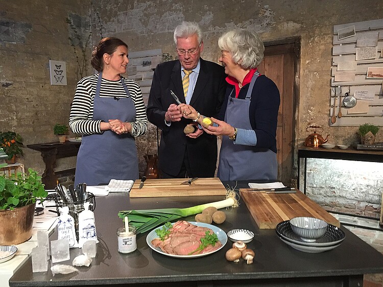 Drei Personen stehen in einer historisch eingerichteten Küche hinter einem Tisch und sprechen miteinander.