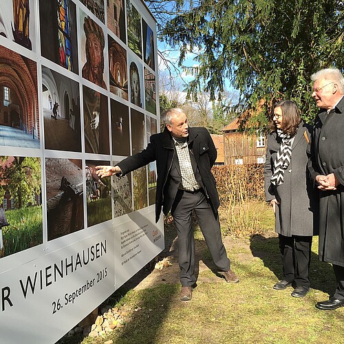 Drei Personen stehen vor einer Wand, die mit Fotos bedruckt ist.