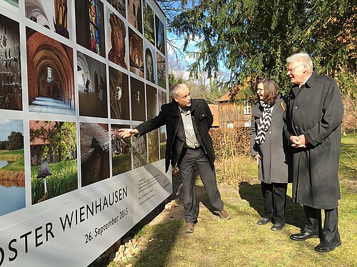 Drei Personen stehen vor einer Wand, die mit Fotos bedruckt ist.