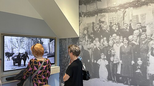 Zwei Frauen, die von hinten zu sehen sind, schauen sich eine Ausstellung mit historischem Filmmaterial und Fotos an