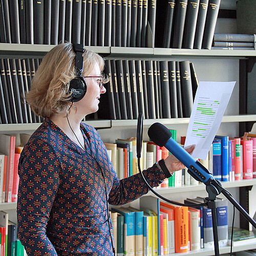 Frau mit Kopfhörern steht vor einem Mikrofon und nimmt einen Text auf, im Hintergrund eine Bücherwand