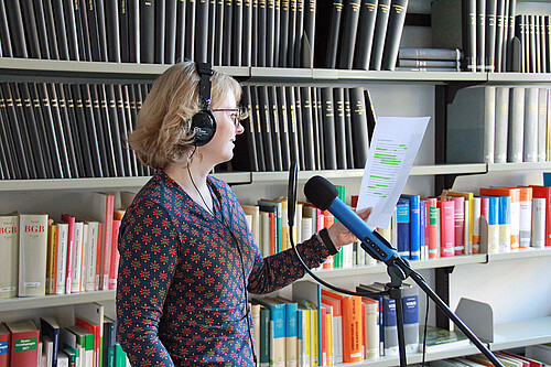 Frau mit Kopfhörern steht vor einem Mikrofon und nimmt einen Text auf, im Hintergrund eine Bücherwand