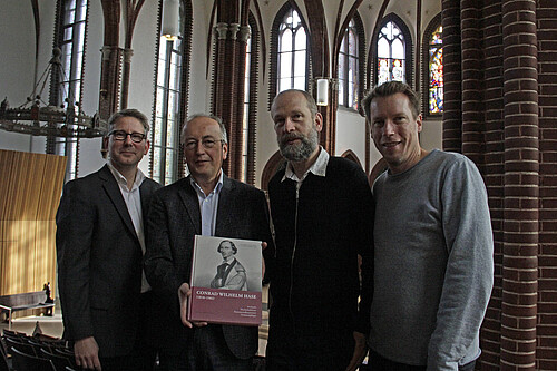 Vier Männer stehen nebeneinander in der Christuskirche in Hannover, einer von ihnen hält ein Buch in den Händen.