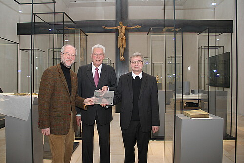 Drei Männer, darunter Klosterkammer-Präsident Hans-Christian Biallas, stehen in einem Raum mit Glasvitrinen und halten ein Schild.