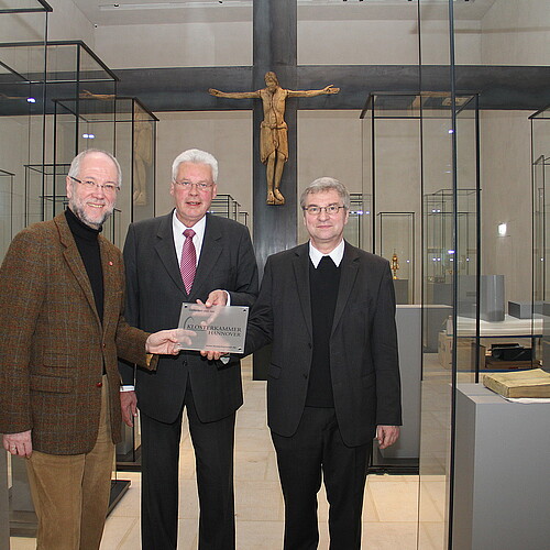 Drei Männer, darunter Klosterkammer-Präsident Hans-Christian Biallas, stehen in einem Raum mit Glasvitrinen und halten ein Schild.