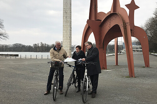Zwei Männer und eine Frau stehen jeweils mit einem Fahrrad in der Hand oder darauf sitzend am Nordufer des Maschsees.