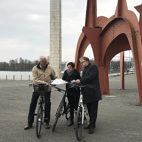 Zwei Männer und eine Frau stehen jeweils mit einem Fahrrad in der Hand oder darauf sitzend am Nordufer des Maschsees.