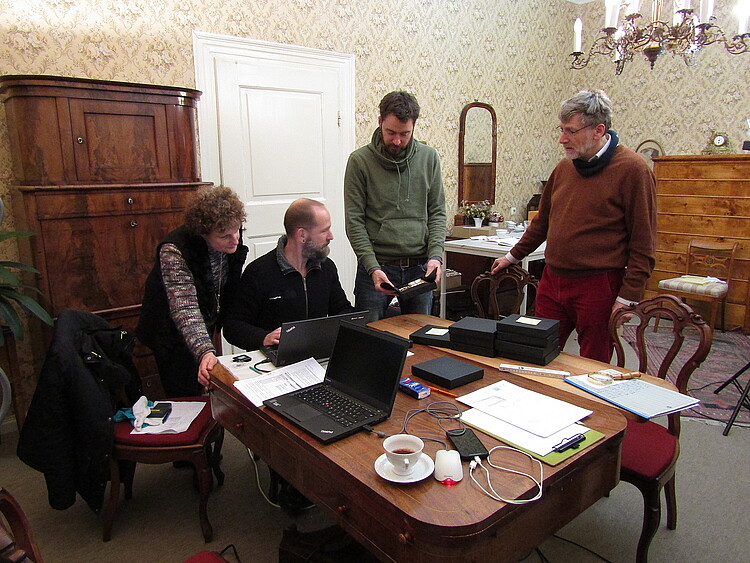 Vier Personen sind um einen Tisch gruppiert, auf dem ein Laptop und andere technische Gerätschaften stehen.