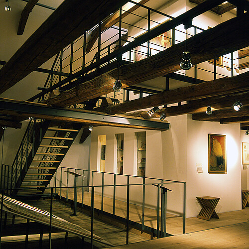 Blick in einen Raum mit mehreren Ebenen, die über eine Treppe verbunden sind. An den Wänden hängen Bilder.