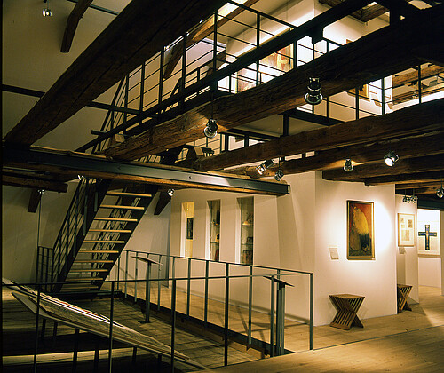 Blick in einen Raum mit mehreren Ebenen, die über eine Treppe verbunden sind. An den Wänden hängen Bilder.