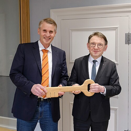 Zwei Männer halten mit großem Holzschlüssel in den Händen, vor einer weißen Holztür