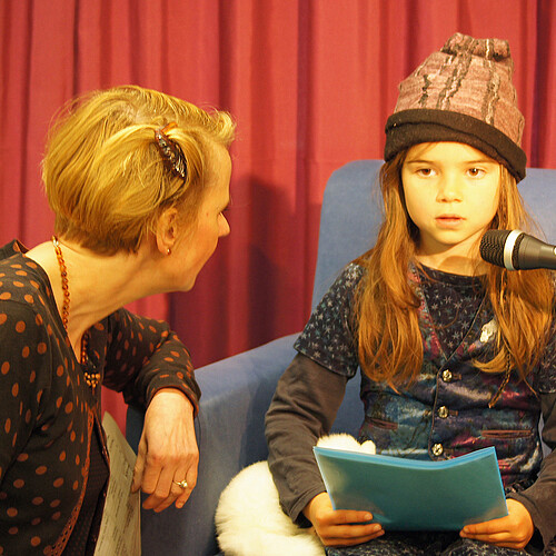 Ein Mädchen sitzt vor einem Mikrophon und liest aus einem Buch vor, neben ihr ist eine Frau zu sehen.