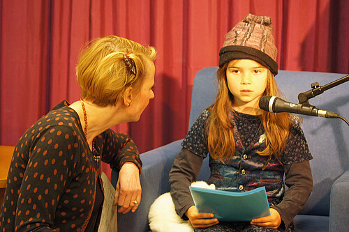 Ein Mädchen sitzt vor einem Mikrophon und liest aus einem Buch vor, neben ihr ist eine Frau zu sehen.
