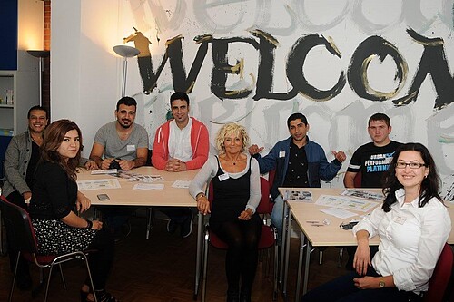 Eine Gruppe junger Männer und Frauen sitzt in einem Raum an Tischen.