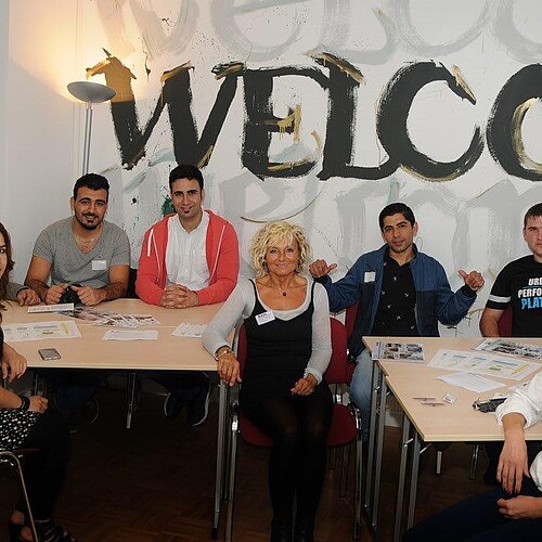 Eine Gruppe junger Männer und Frauen sitzt in einem Raum an Tischen.