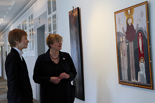 Zwei Frauen schauen auf ein Bild, das an einer Wand im Kreuzgang hängt.