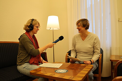 Zwei Frauen sitzen an einem Tisch. Eine der beiden hält ein Mikrofon und trägt Kopfhörer, die andere spricht und hält Blätter in den Händen.