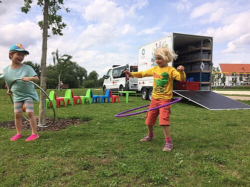 Zwei Kinder spielen mit Hula-Hoop-Reifen in einem Park, im Hintergrund ist ein Auto mit einem Anhänger zu sehen.