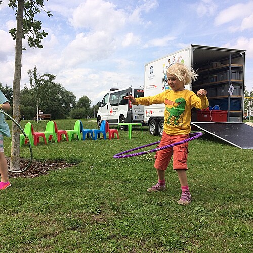 Zwei Kinder spielen mit Hula-Hoop-Reifen in einem Park, im Hintergrund ist ein Auto mit einem Anhänger zu sehen.
