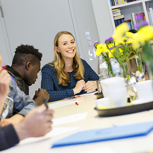 Eine junge Frau sitzt an einem Tisch gemeinsam mit zugewanderten Jugendlichen.