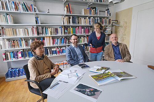Drei Personen sitzen an einem Tisch, eine Person steht mit einem Mikrofon dazwischen.