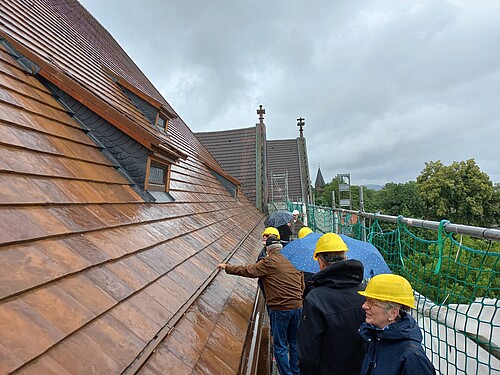 Mehrere Personen mit Bauhelmen und Regenschirmen bei Regen auf einem Baugerüst vor einem frisch gedeckten Kirchendach.